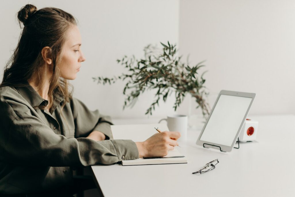 woman writing down notes while looking at tablet